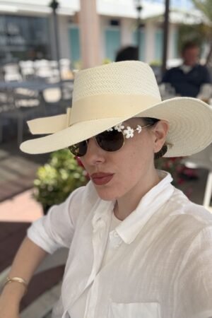 Primer plano de una mujer luciendo un sombrero de paja trenzado ajustable con una elegante cinta en color crudo, combinado con una blusa de lino blanca de la marca OH.LA.
