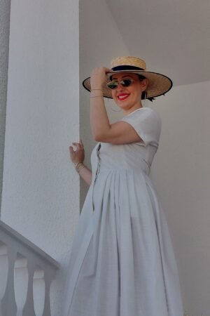 Mujer sonriente en una escalera exterior luciendo una pamela de ala ancha de paja natural con detalles en azul marino, combinada con un vestido blanco de la marca OH.LA.