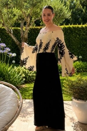 Mujer posando en un jardín vistiendo el Top Lexie, una prenda off-the-shoulder en color crema con cuerpo elástico nido de abeja y mangas acampanadas con bordados geométricos en negro, combinada con pantalones negros anchos. Se muestran vistas frontal, lateral y trasera.