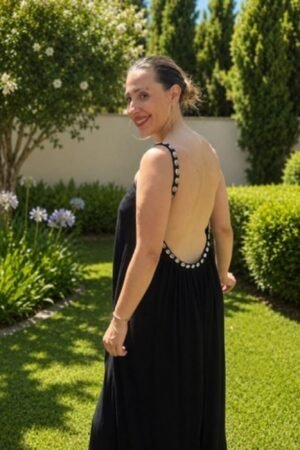 Mujer de espaldas luciendo un vestido largo negro con un gran escote trasero bordeado de conchas blancas, en un jardín soleado.