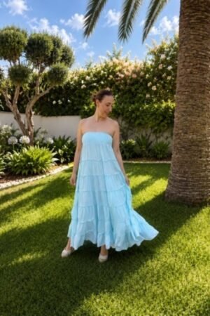 Una mujer posa de frente con un vestido largo de volantes color aqua y escote palabra de honor, mirando hacia abajo en un jardín con palmeras.